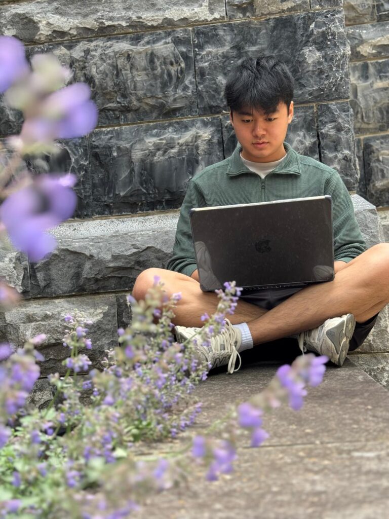 A senior elemage named Hinek is working on a project while sitting criss-cross infront of a stone wall near lavender flowers
