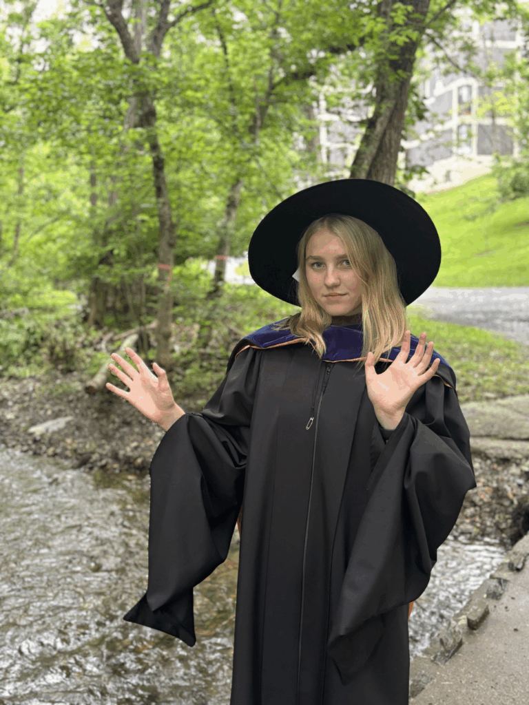 Woman standing in front of a creek wearing black robes and witch hat while holding hands in air