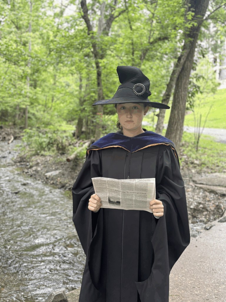 Woman standing in front of creek dressed in black cloak and black witch hat holding a newspaper