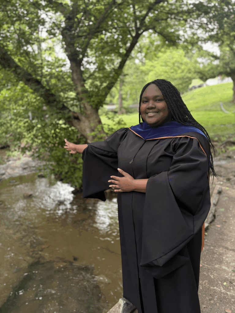 Woman standing in front of creek wearing black robe and gesturing towards stream
