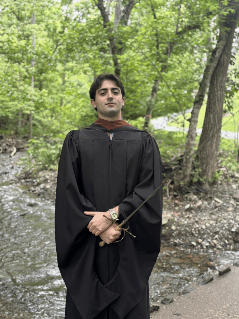 Man standing in front of a creek wearing black cloak while holding a sword.