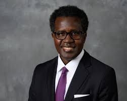 A black man in a suit smiling with a purple tie on